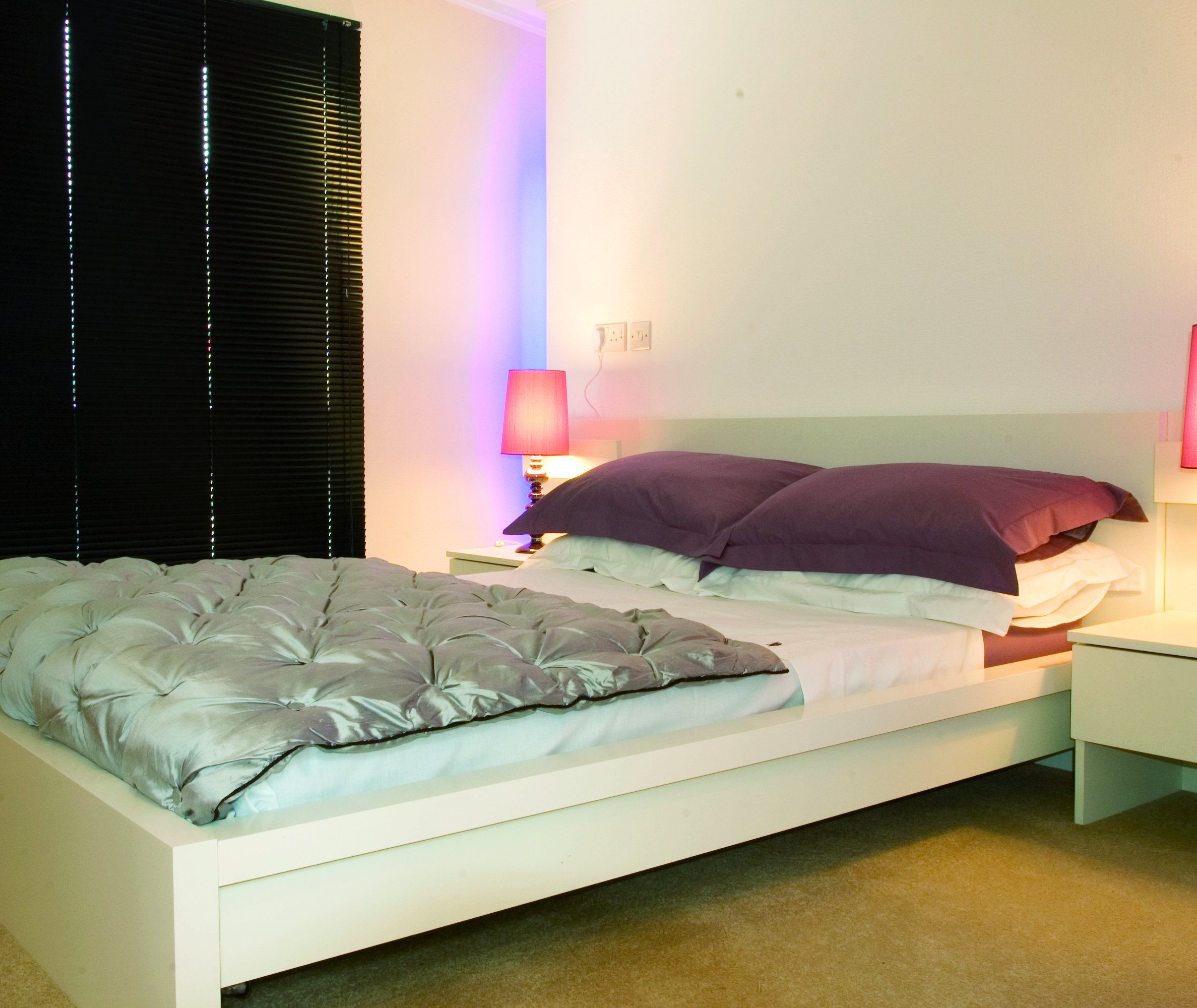 Modern master bedroom with white bed frame, grey quilt, dark pillows and pink bedside lamps beside black blinds