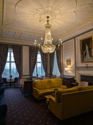 Elegant lounge with yellow sofas, large chandelier, ornate ceiling, and tall windows with drapes