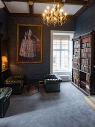 Elegant historic library room with ornate fireplace, classical portrait, shelves of old books, and green leather chairs