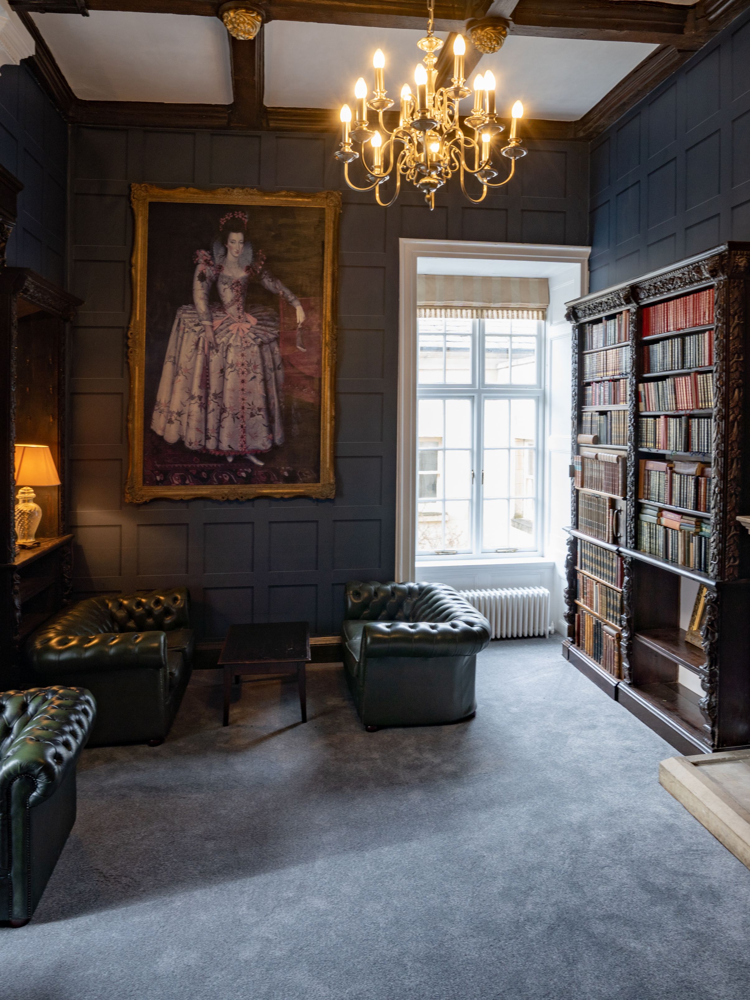 Elegant historic library room with ornate fireplace, classical portrait, shelves of old books, and green leather chairs
