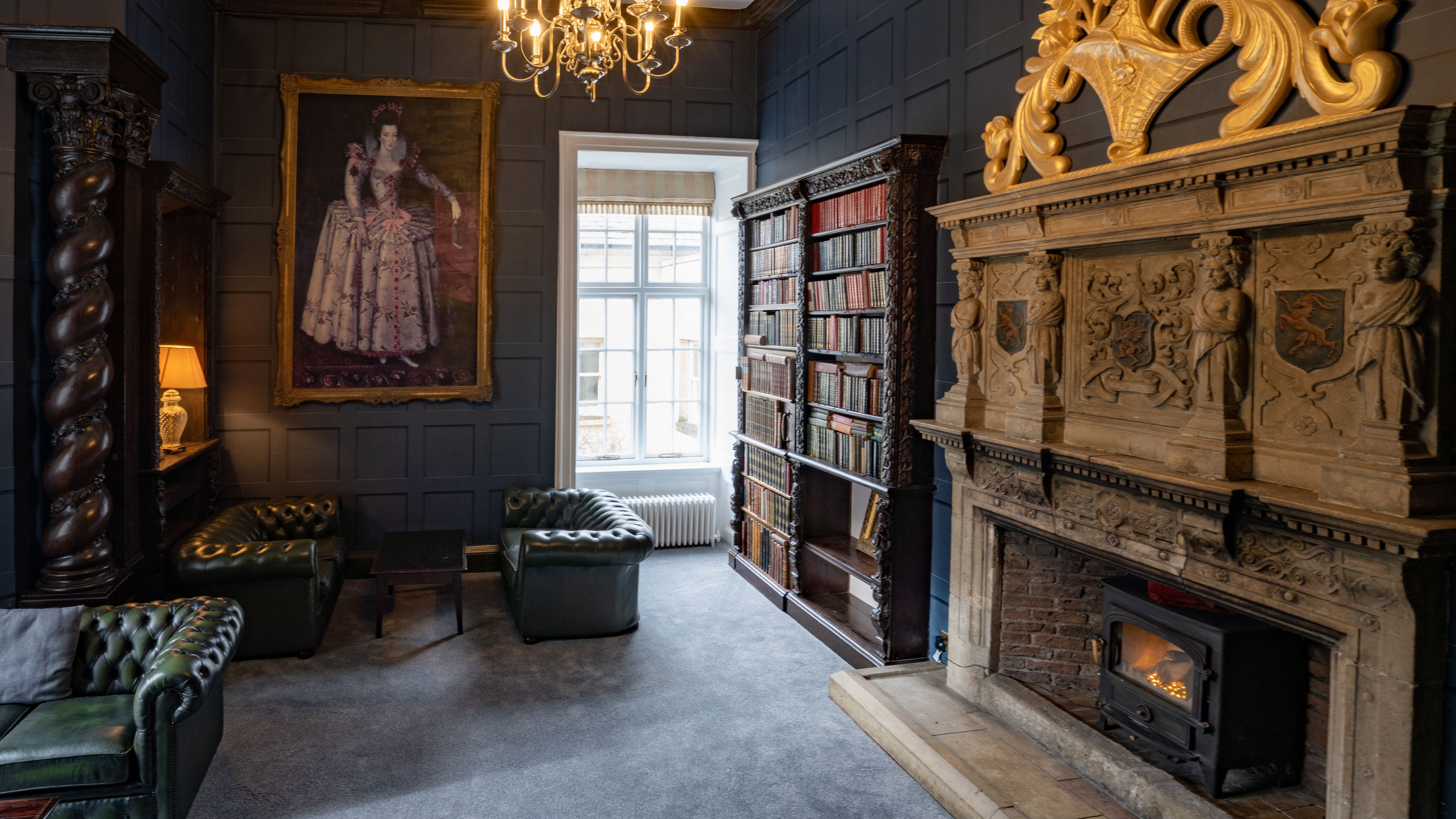 Elegant historic library room with ornate fireplace, classical portrait, shelves of old books, and green leather chairs