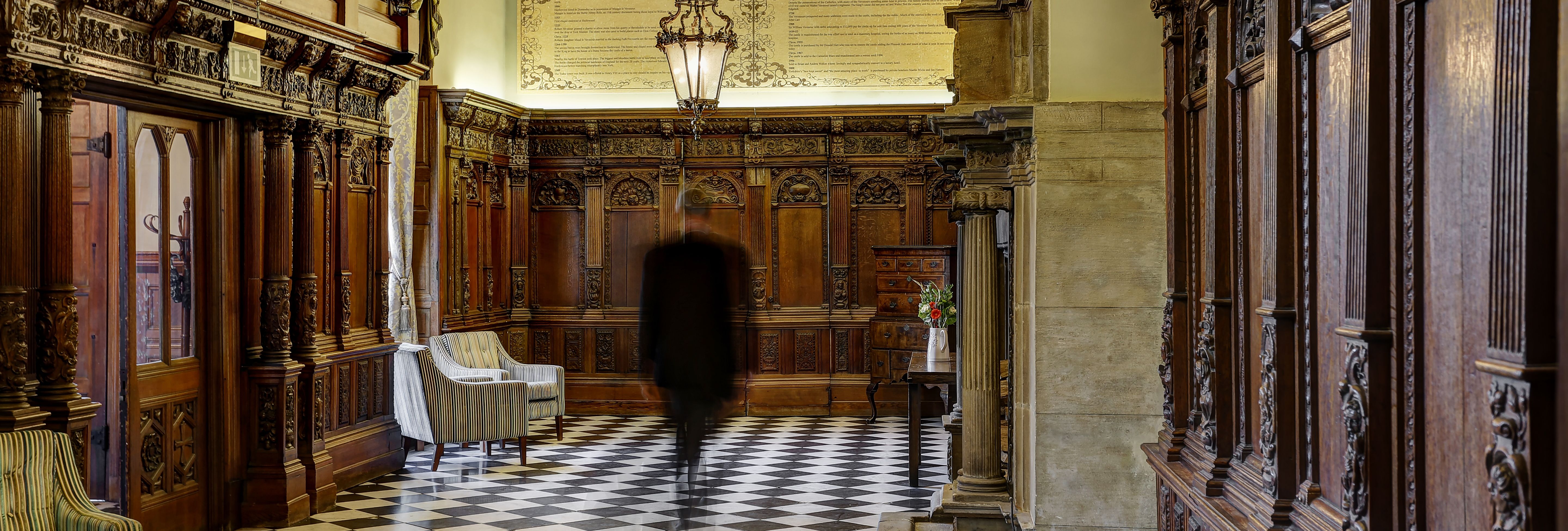 Historic hotel hallway with ornate wood paneling, checkered floor, and elegant lighting