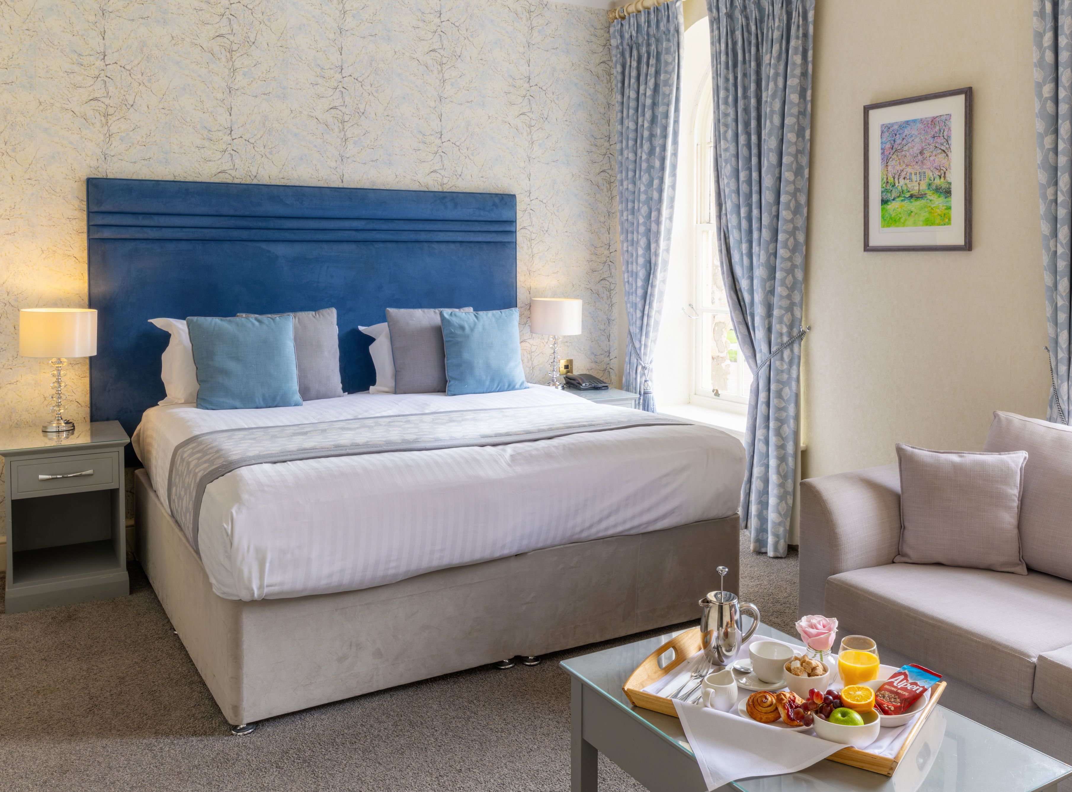 Elegant hotel room with a large double bed, blue headboard, cozy sofa, and breakfast tray on a glass coffee table.