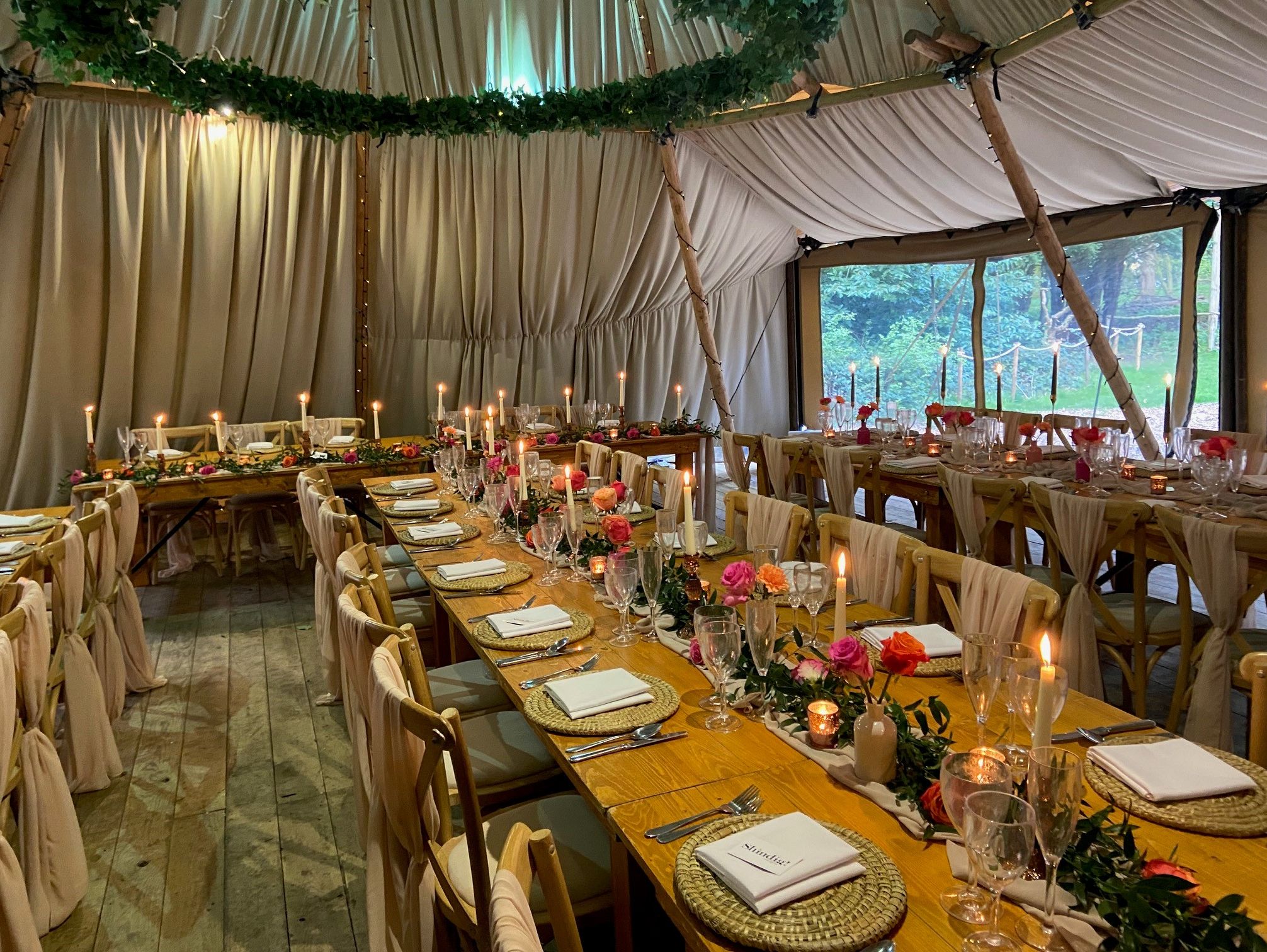 Decorated interior of a tipi set up for an event with long wooden tables, chairs draped in fabric, candles, and floral arrangements.