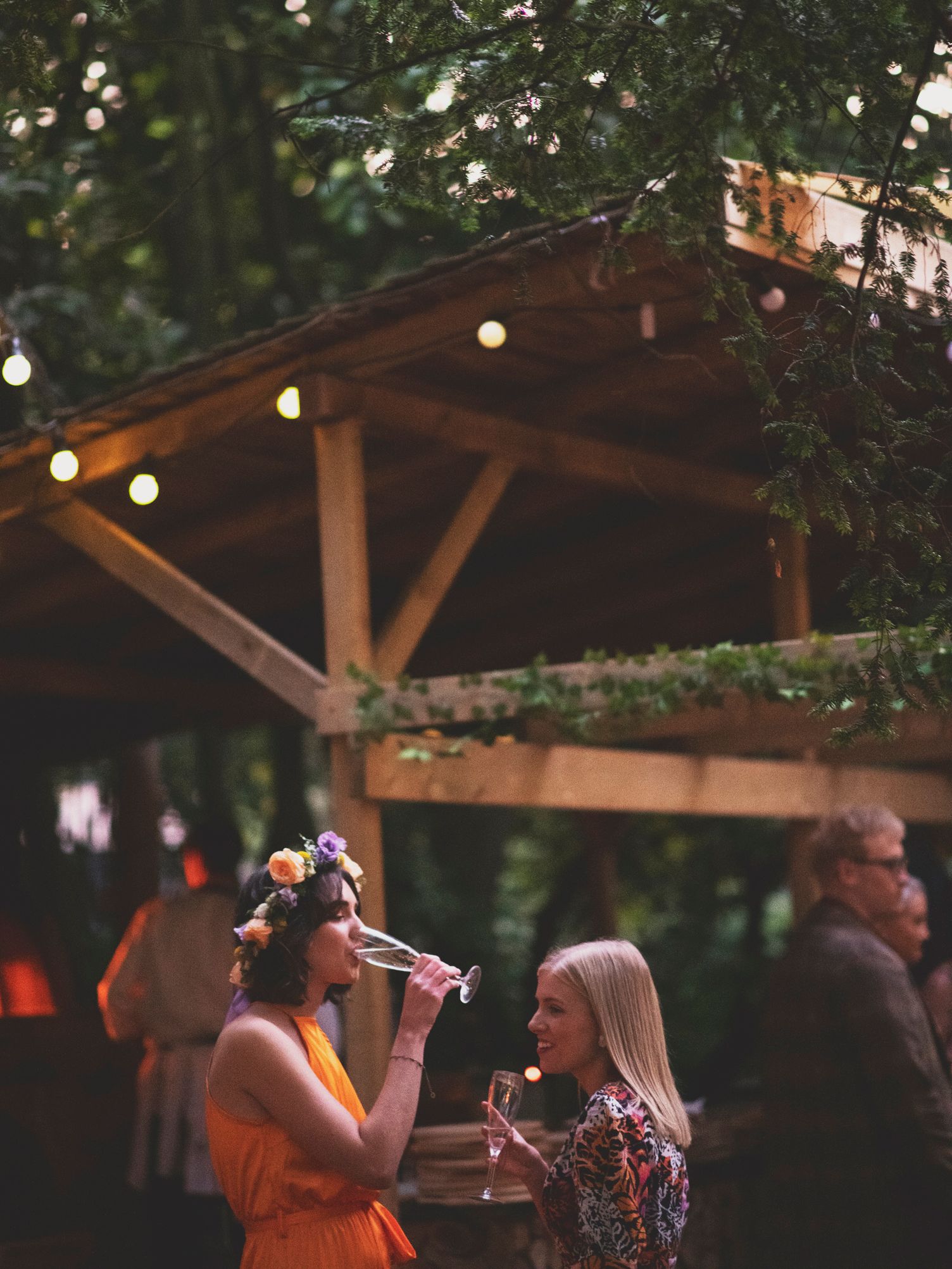 Two women enjoying drinks at an outdoor evening reception under string lights.