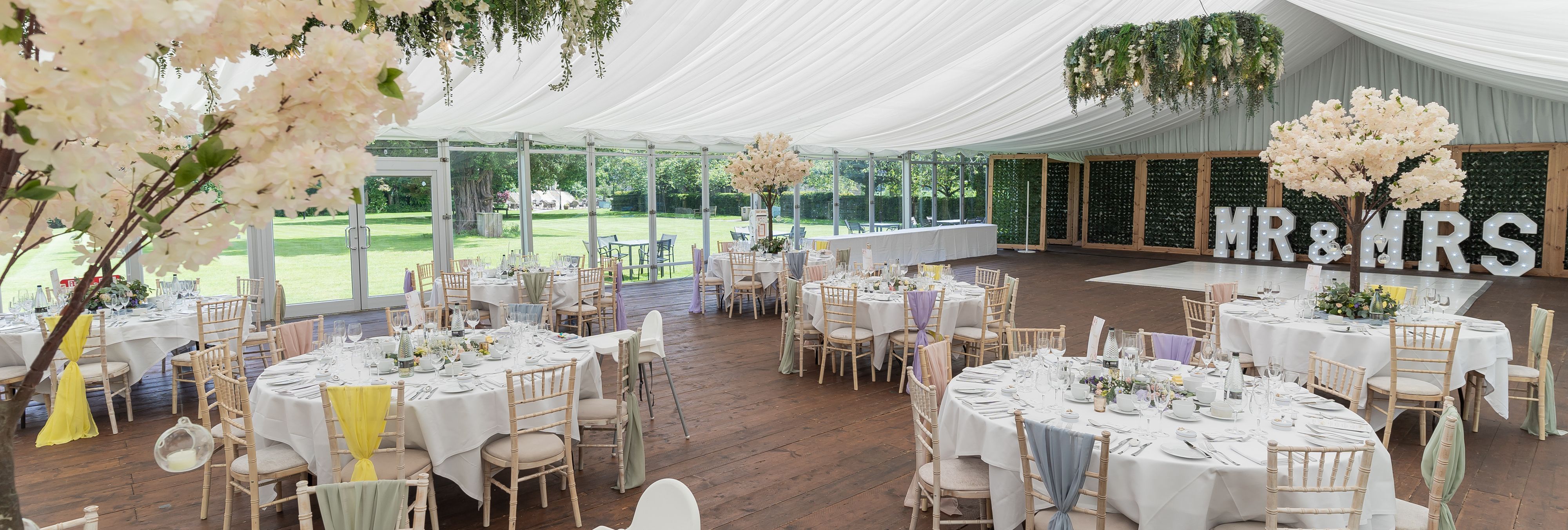 Elegant wedding reception setup in a marquee with white draping, round tables, pastel decorations, and 'MR & MRS' sign in the background.