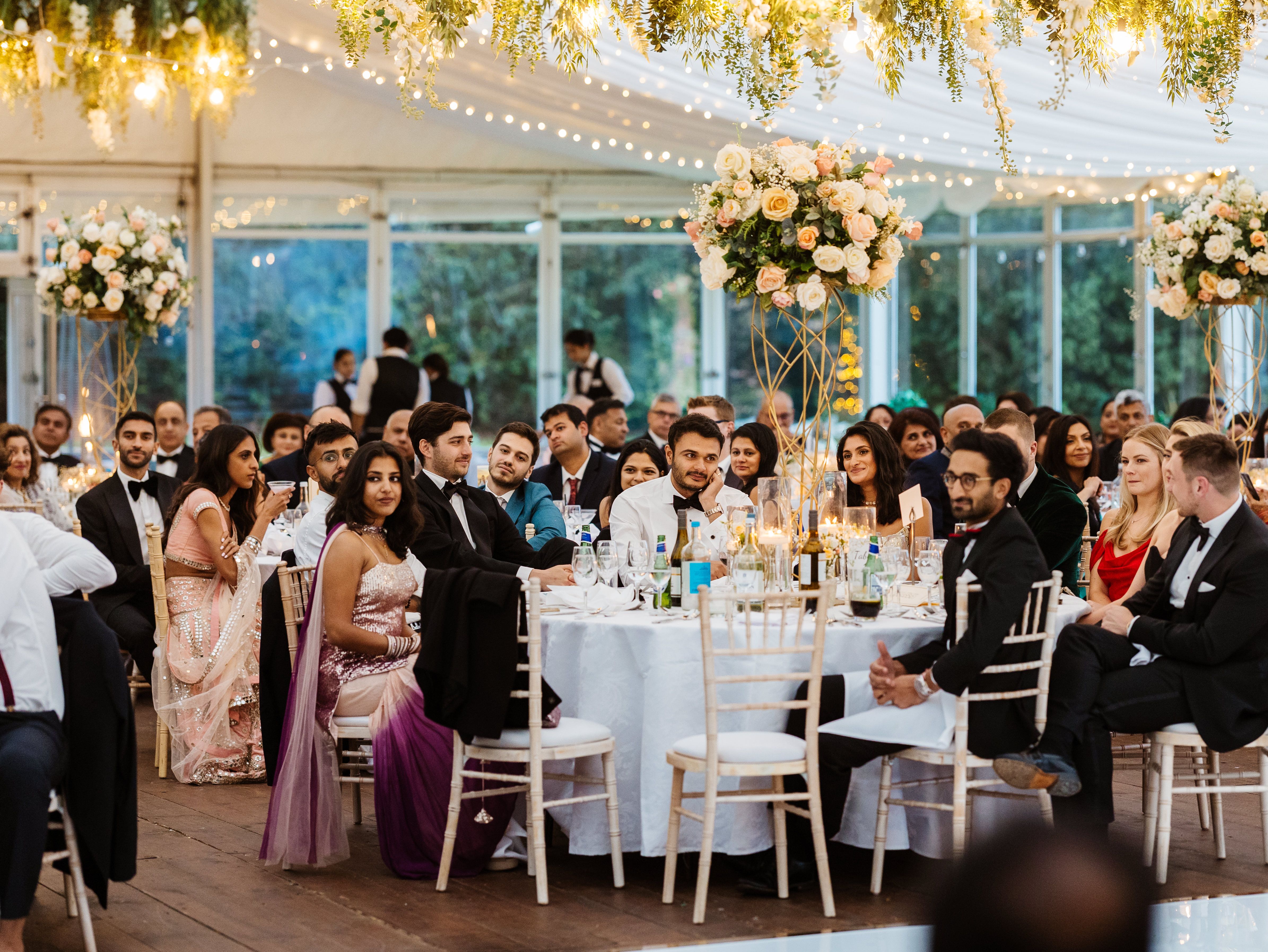 Guests seated at elegantly decorated tables during a wedding reception under a marquee with floral arrangements and hanging greenery.