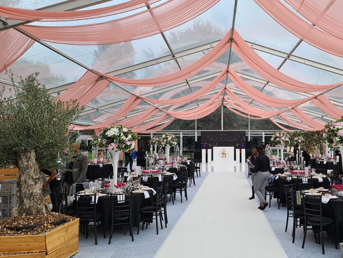 Elegant wedding reception hall with pink draped ceiling, black chairs, round tables with floral centerpieces, and a white aisle.