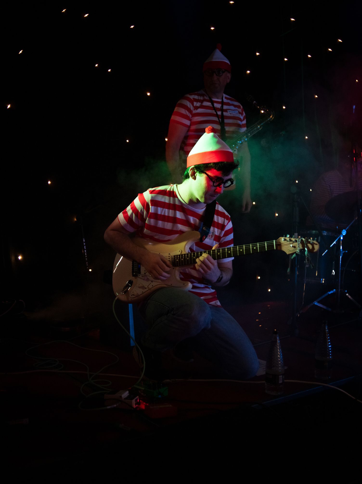 Musicians performing on stage dressed in red and white striped shirts and matching hats, with one playing electric guitar and another on saxophone.