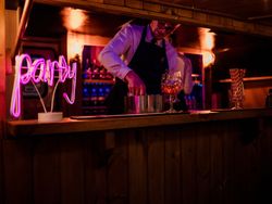 Bartender preparing drinks behind a wooden bar with a neon sign that reads 'party'.