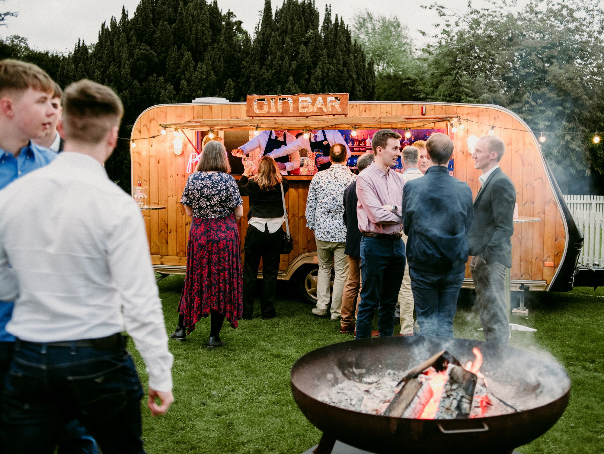 People socializing outside at an evening reception with a gin bar trailer and a fire pit.