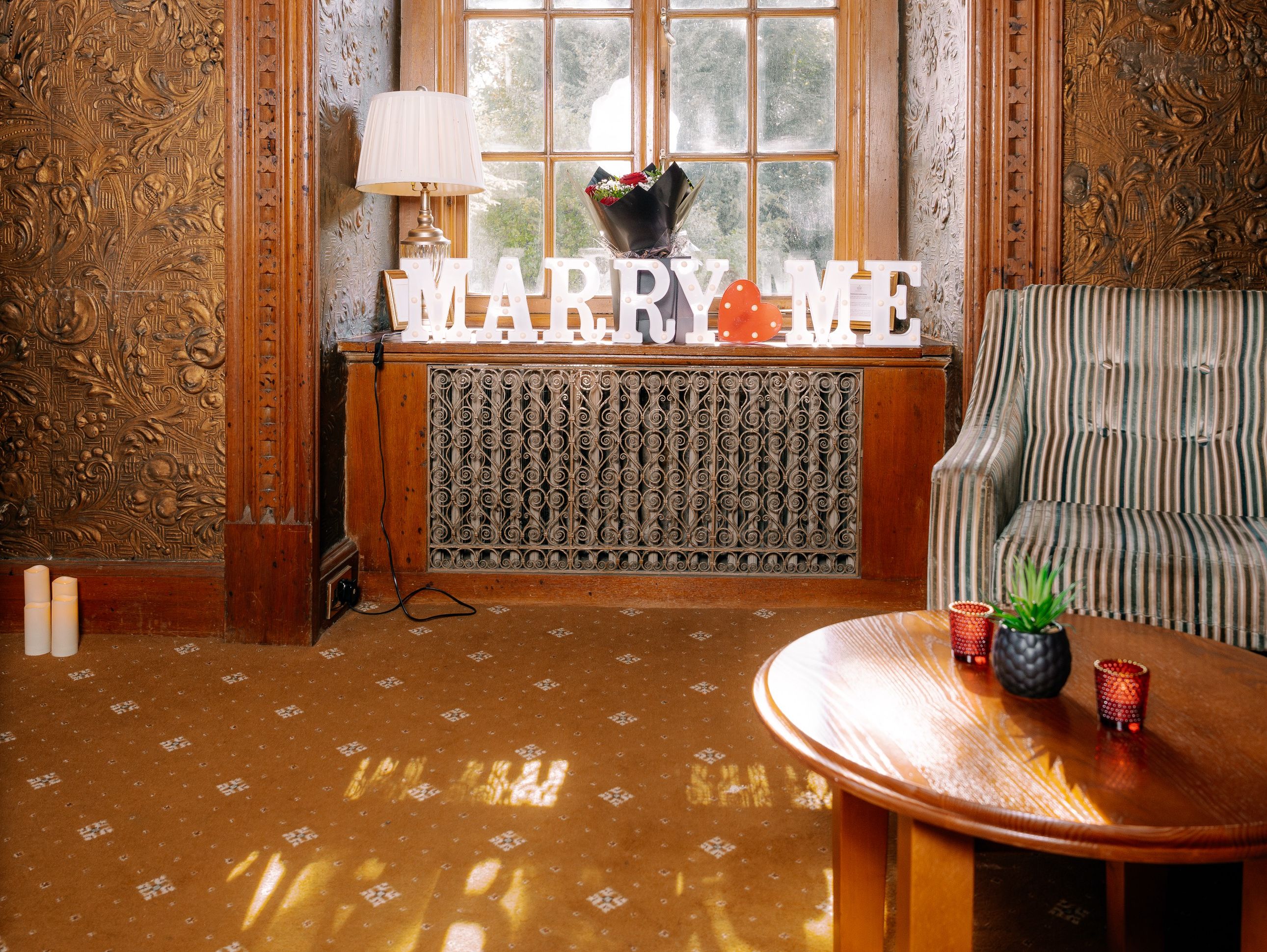 Decorative interior scene featuring a window with a 'MARRY ME' sign and a bouquet in front of it.