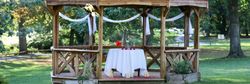 Wooden gazebo decorated with white ribbons and a table set for two with candles and flowers in a green park.