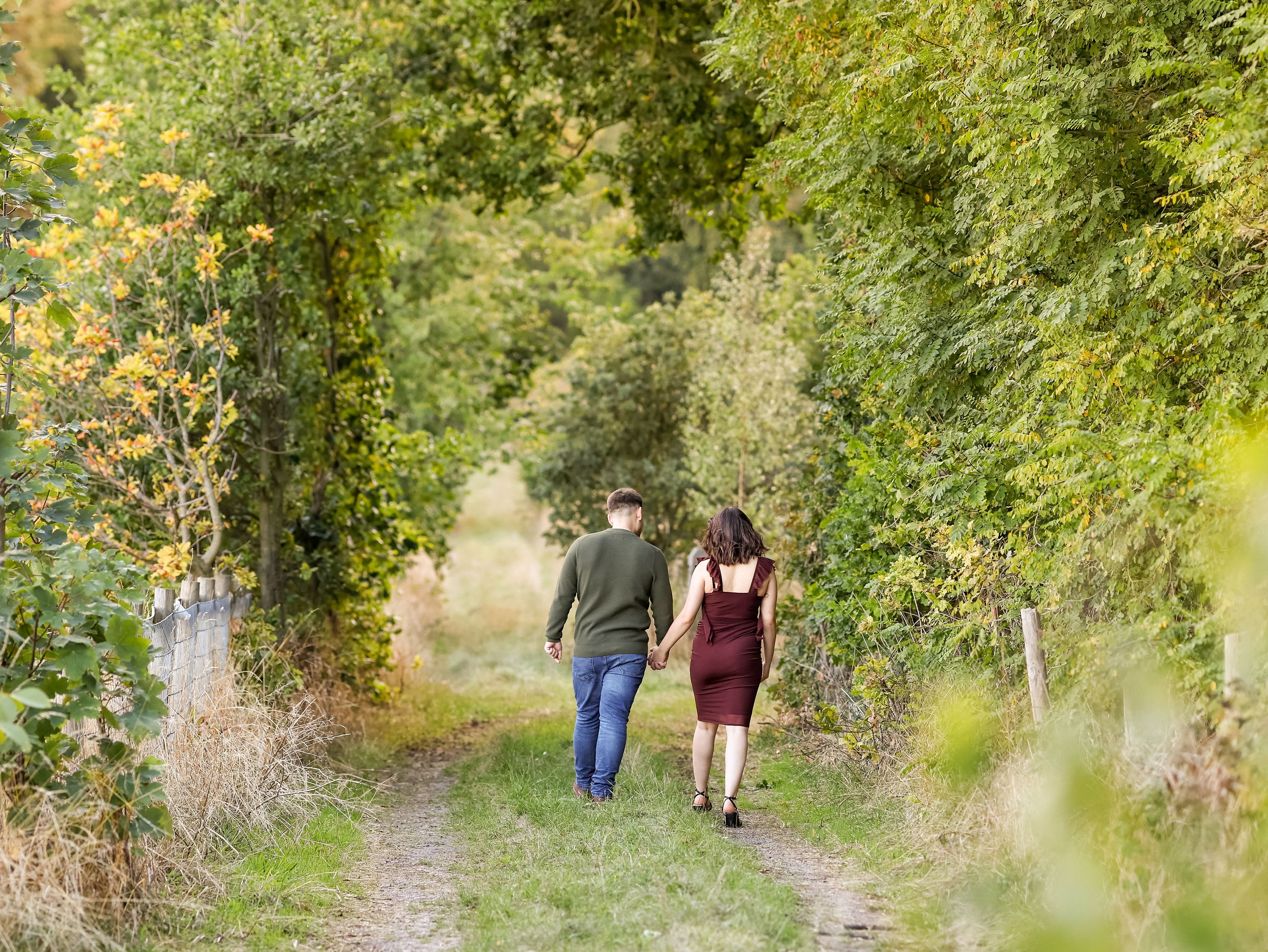A couple holding hands walks down a tree-lined path.