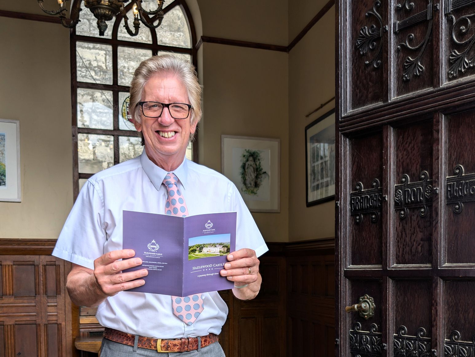 Smiling man wearing glasses and a white shirt with a pink spotted tie stands inside near a wooden door, holding a Hazelwood Castle brochure.