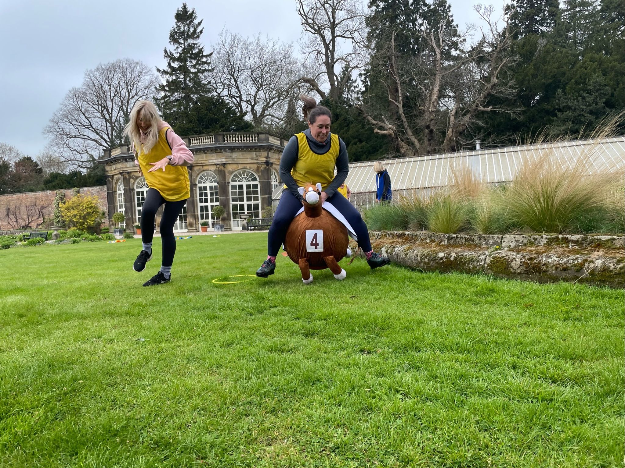 Two women in yellow vests participating in a playful outdoor race, one jumping and one riding a bouncing horse toy.