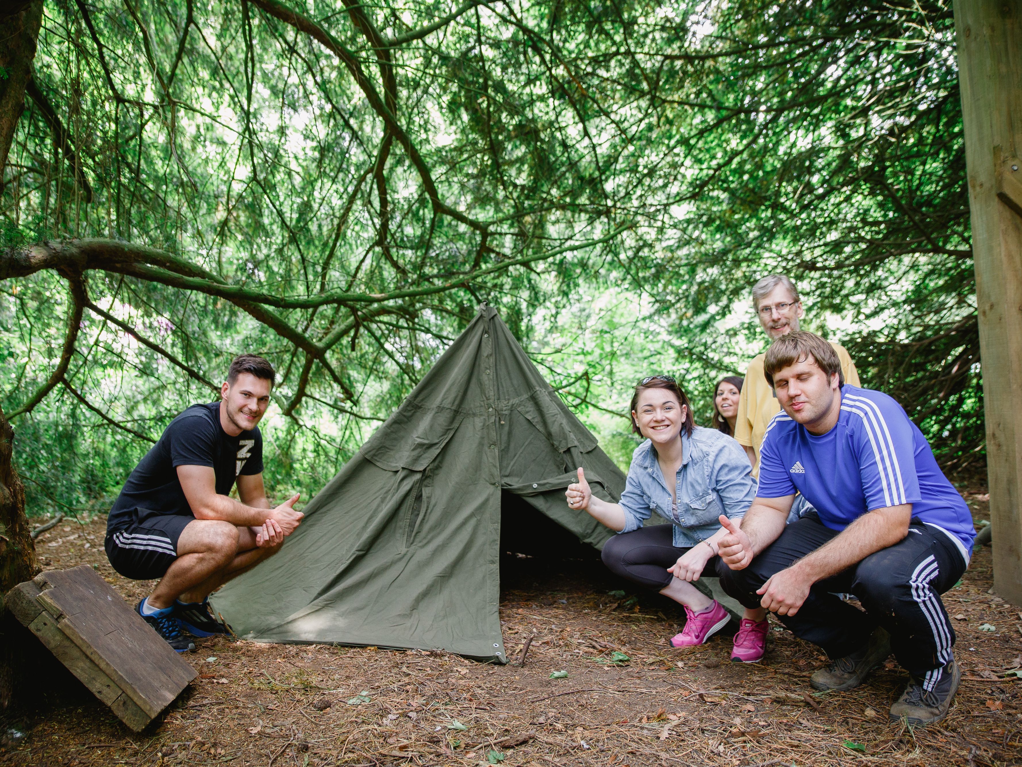Group of people giving thumbs up in front of a green tent in a wooded area.