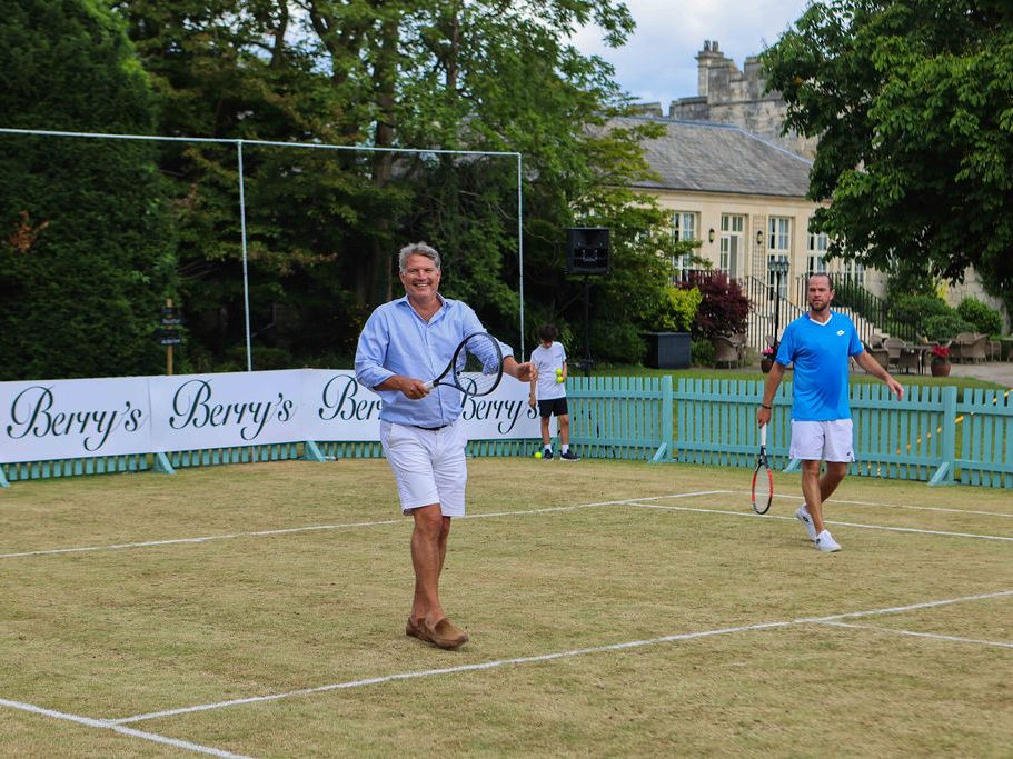 Men playing tennis on an outdoor grass court with a green fence and Berry's advertisement in the background.