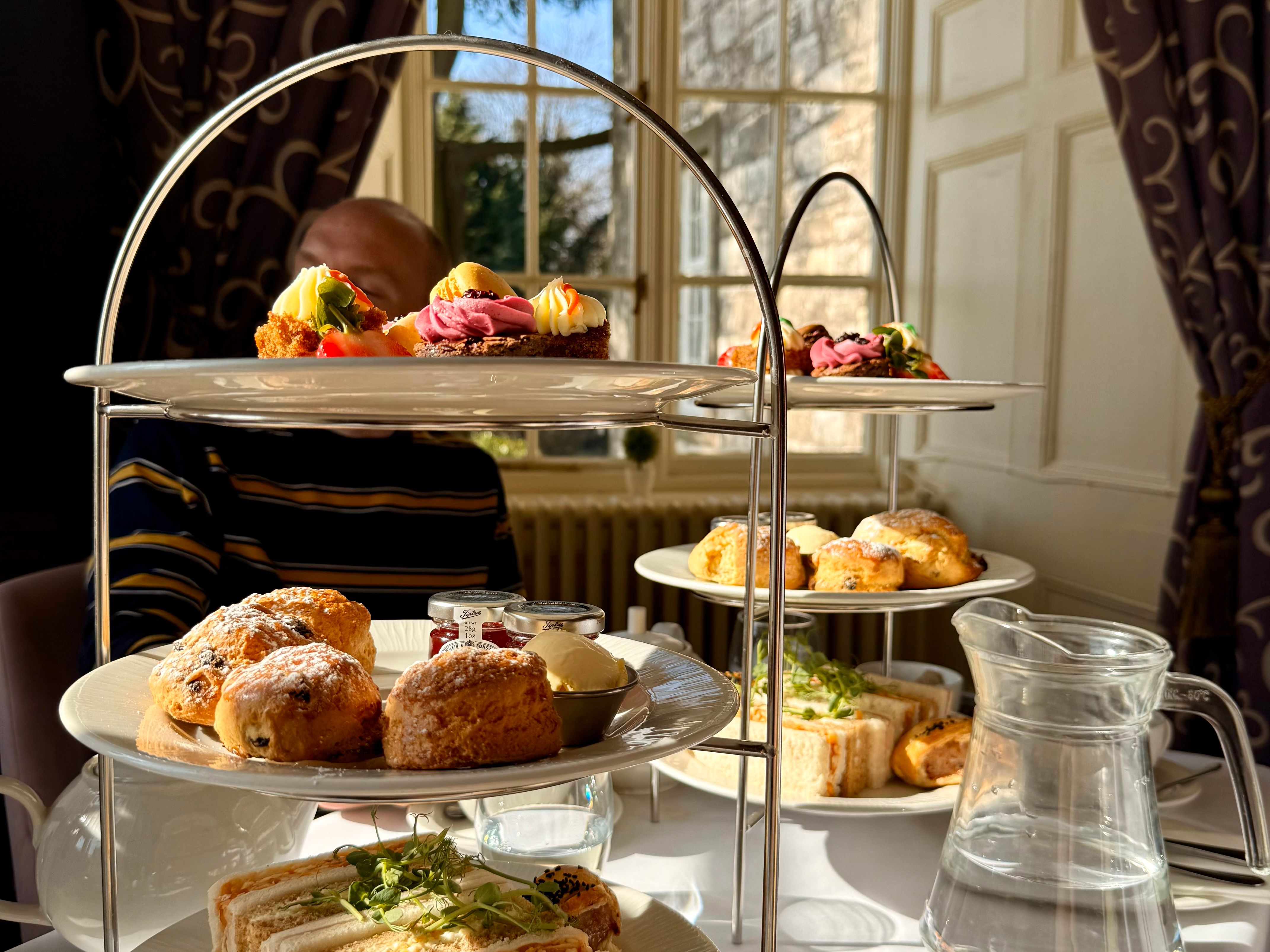 Three-tiered afternoon tea set with sandwiches, scones, pastries, and a cup of tea on a sunlit table by a window
