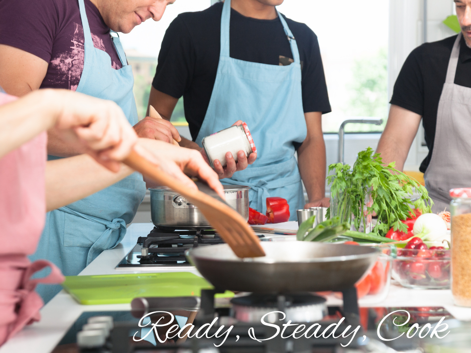 Group of people wearing aprons cooking together in a kitchen with fresh vegetables and herbs on the counter.