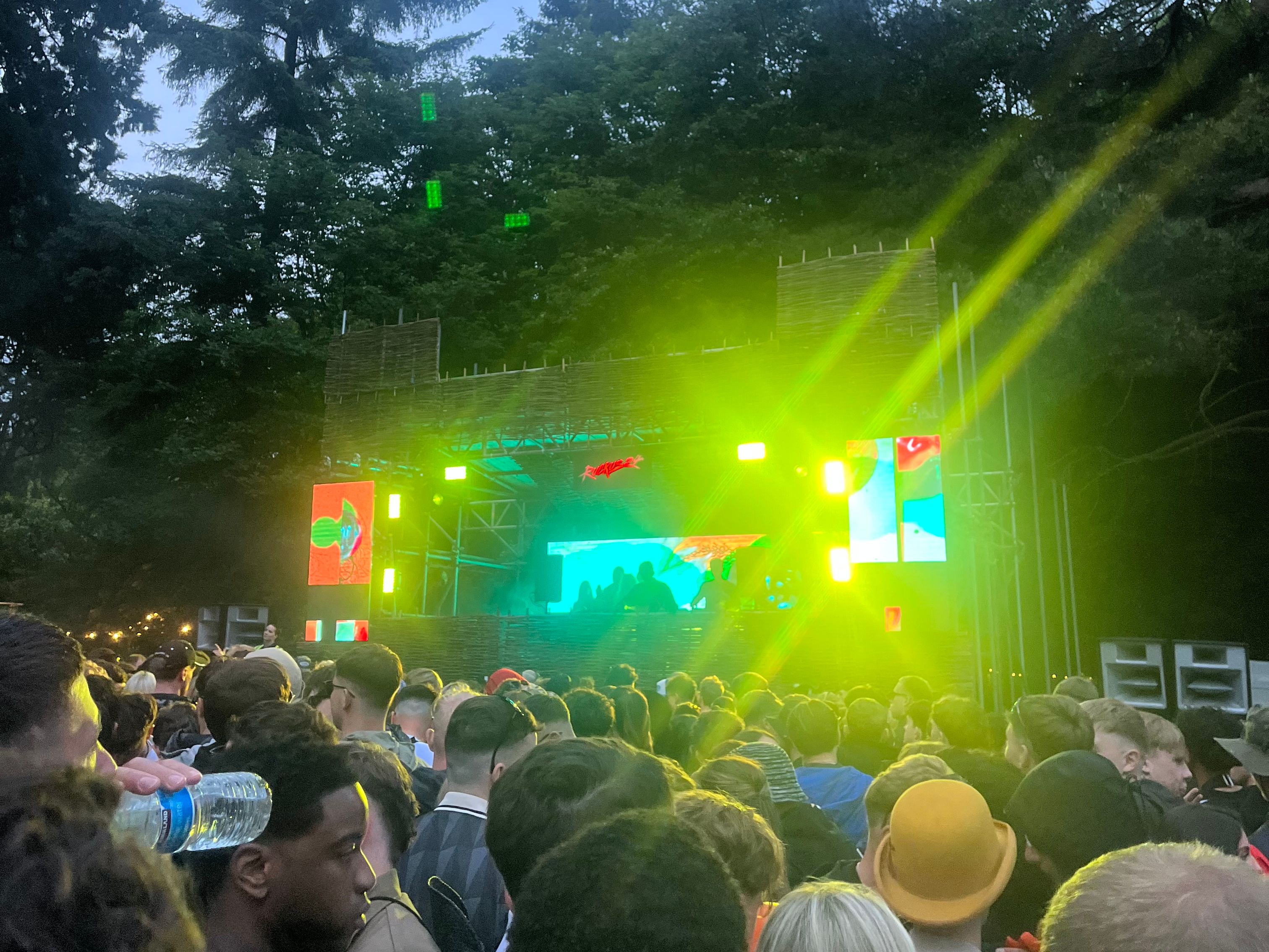 Outdoor music festival with a crowd in front of a stage lit by colorful lights in a forested area.