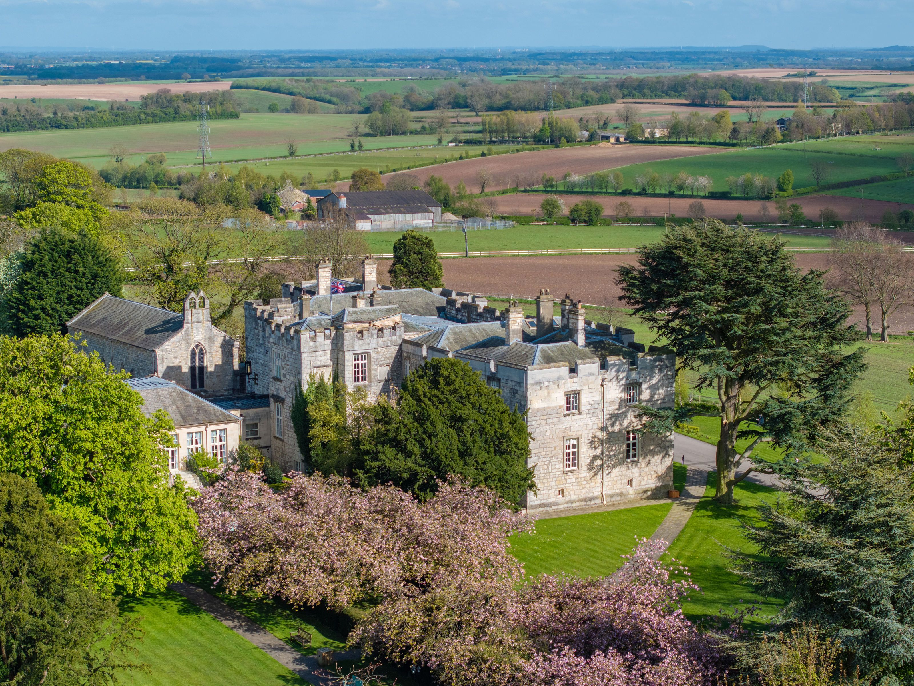 Aerial view of a historic stone manor house surrounded by lush trees and gardens, with open countryside in the background.