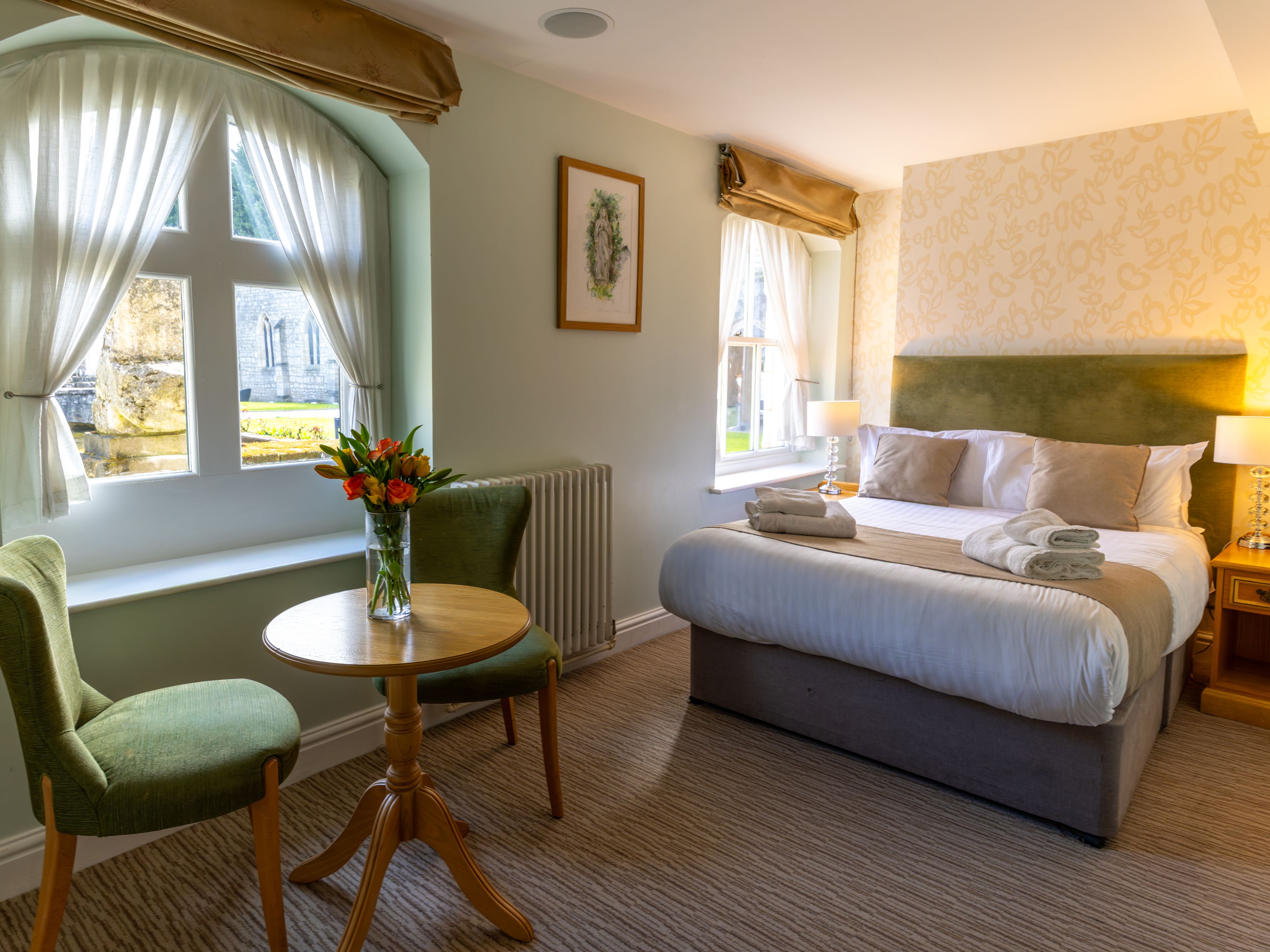 Cozy hotel room with a double bed, two green chairs, a small round wooden table with a vase of flowers, and large windows with sheer curtains.