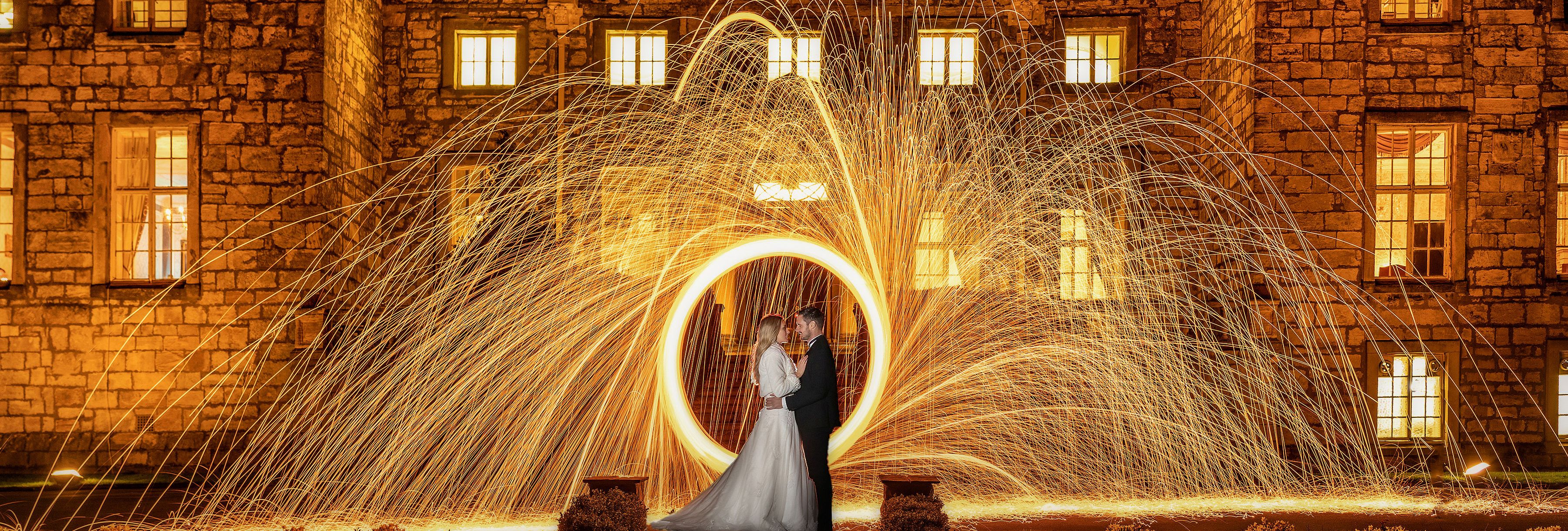 Bride and groom standing in front of a stone building at night, surrounded by a captivating display of swirling golden sparklers.