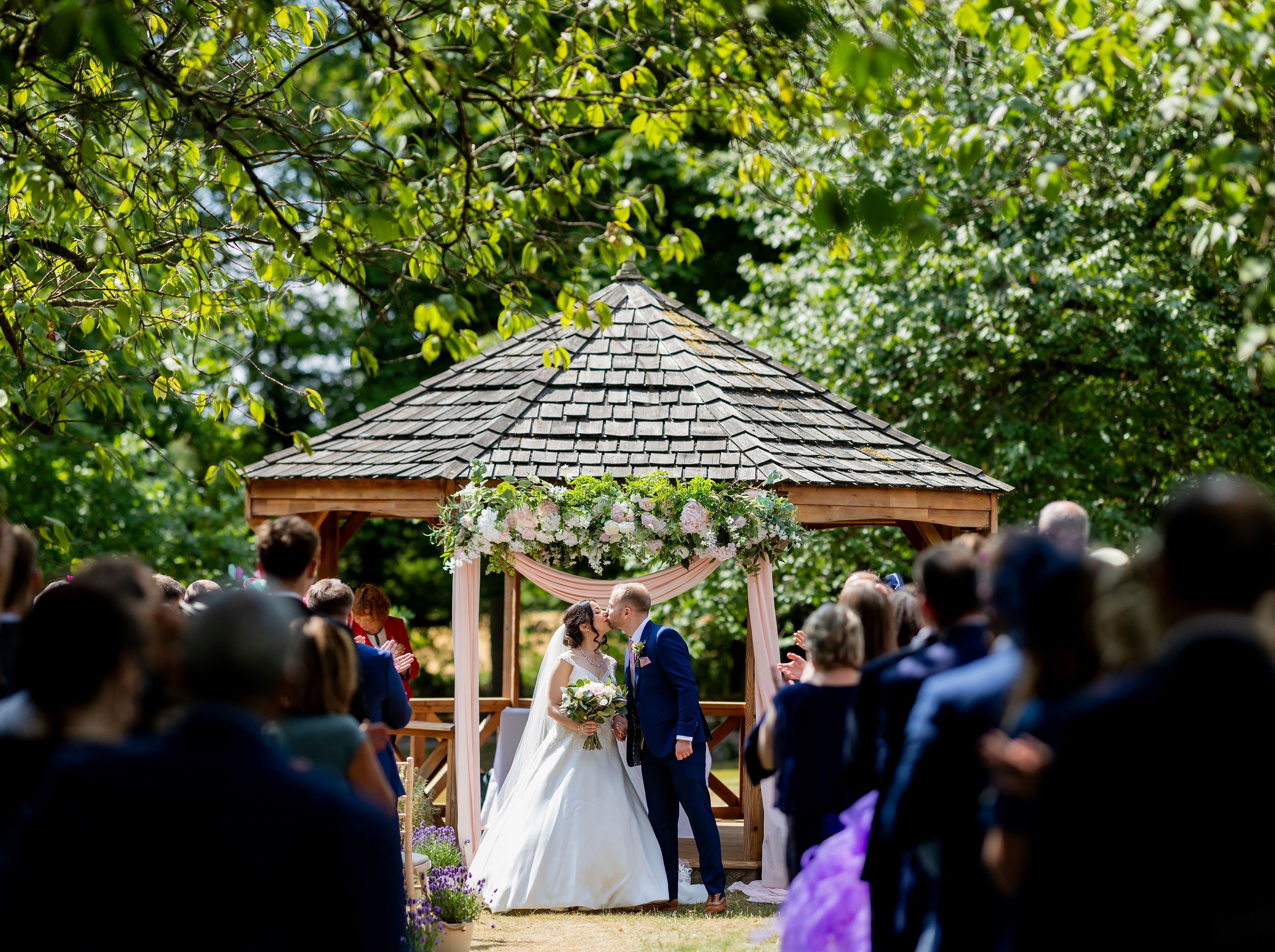 Bride and groom sharing a kiss at an outdoor wedding ceremony under a floral-decorated gazebo with guests in attendance.