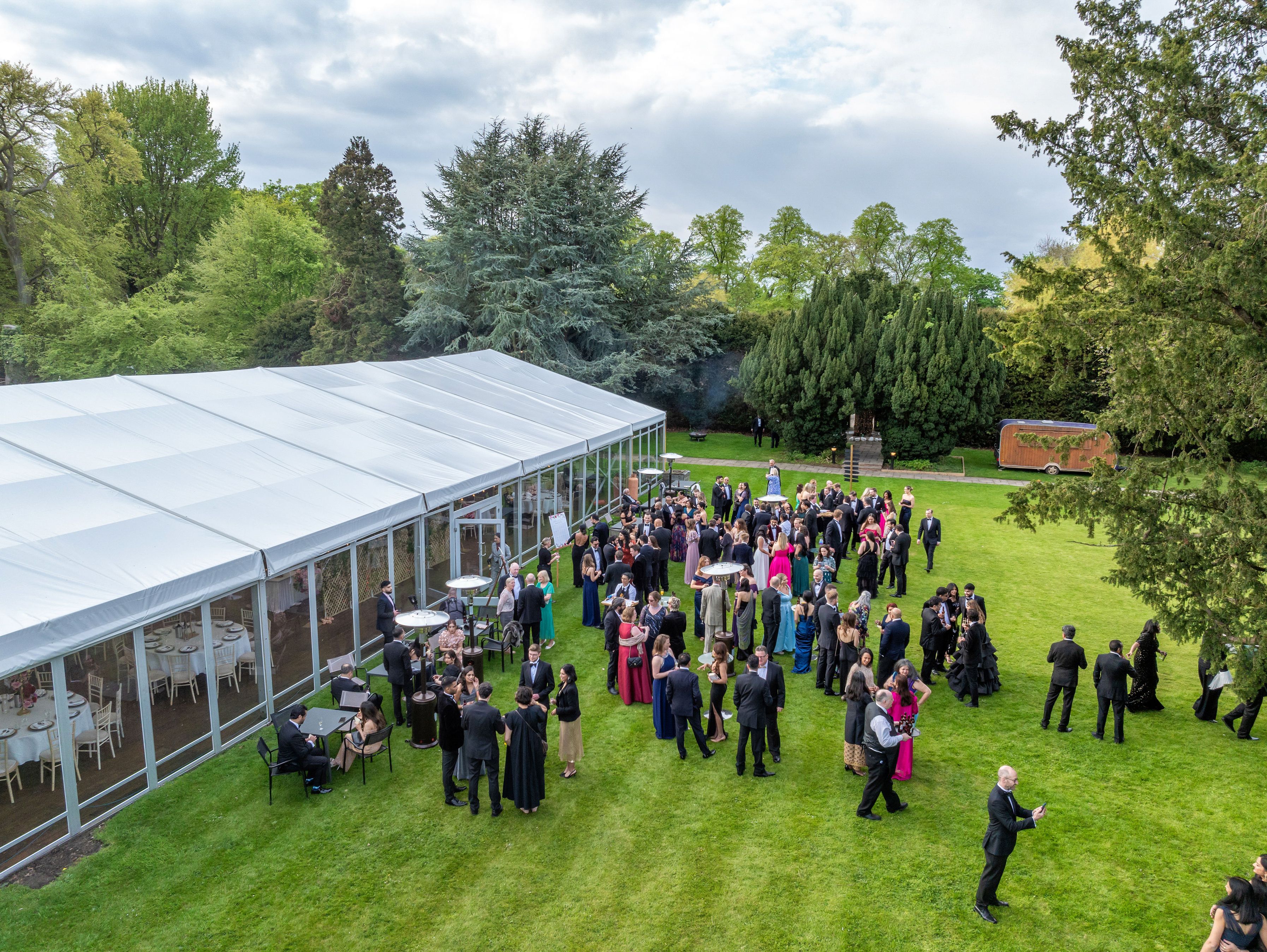 A large group of well-dressed people mingling on a lawn outside a large white event tent, surrounded by trees and greenery.