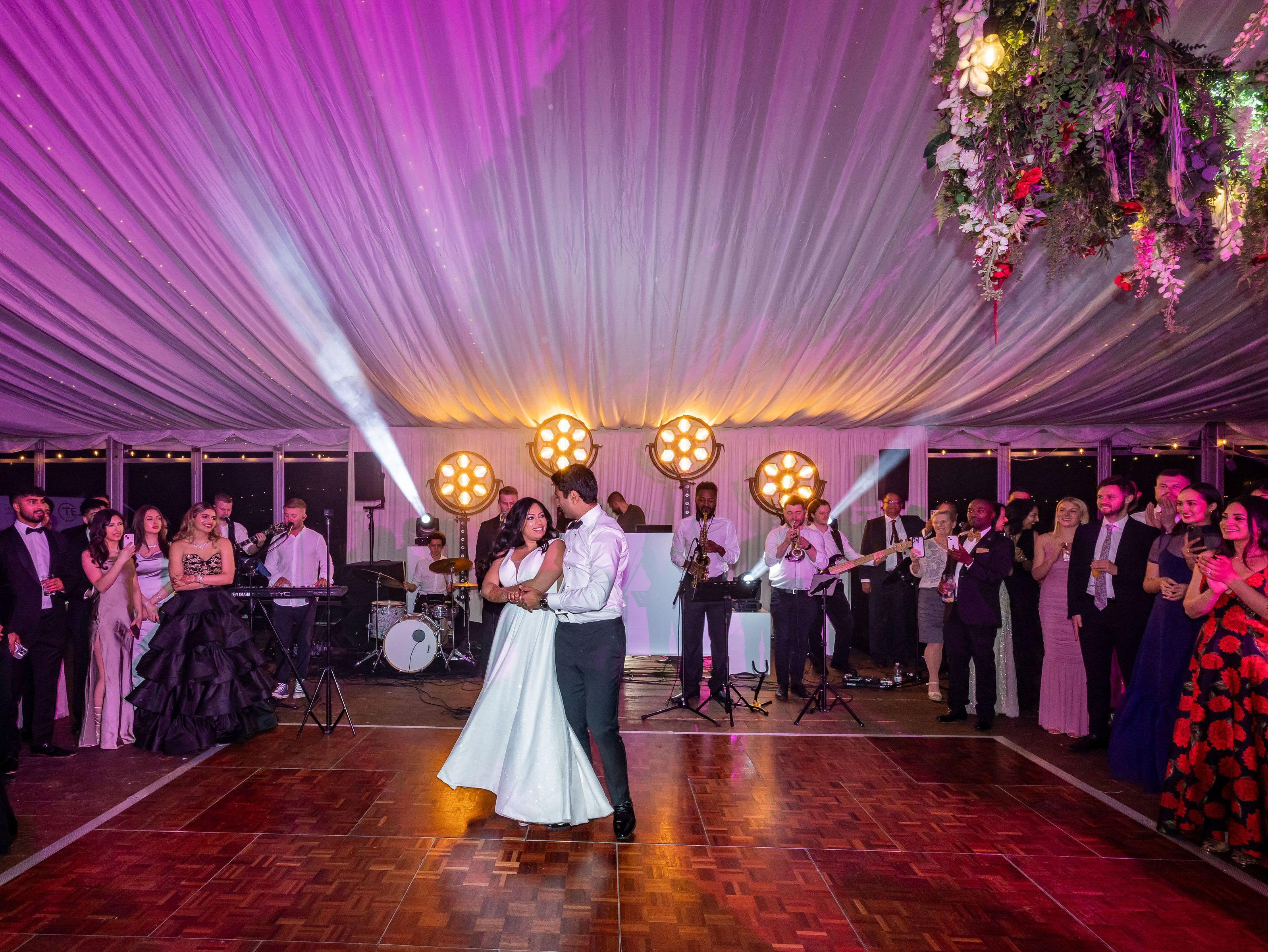 Bride and groom share their first dance at a wedding reception under a decorated tent with live band and guests watching