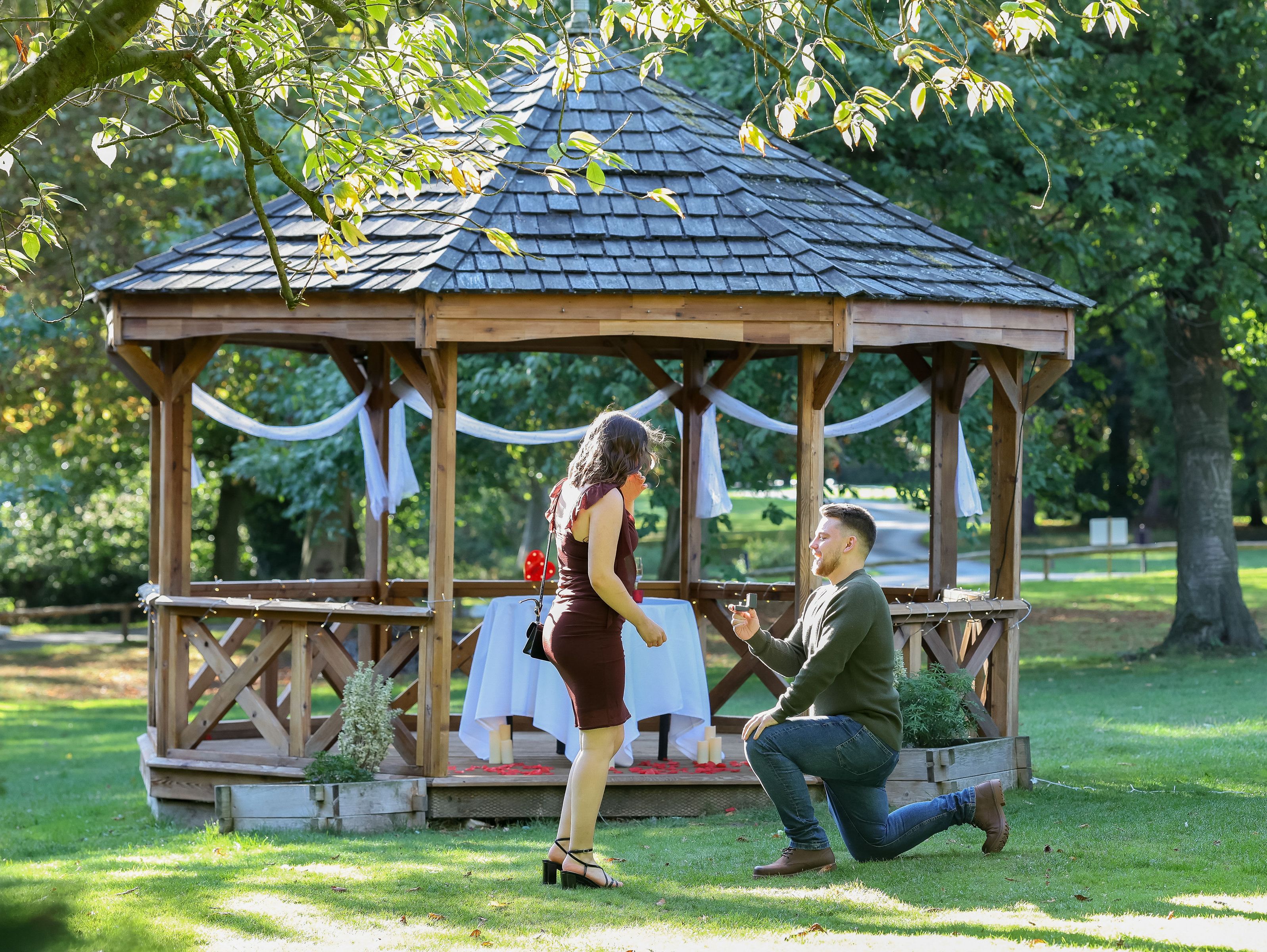 Man proposing to woman in front of a decorated gazebo in a park.