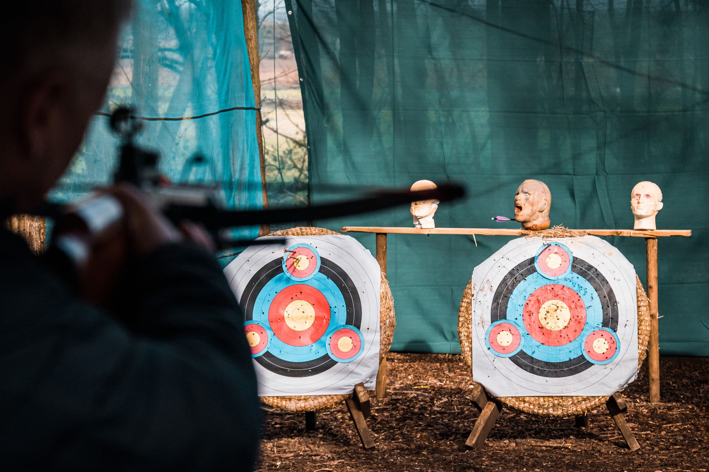 Person aiming a crossbow at two archery targets with mannequin heads placed above them.