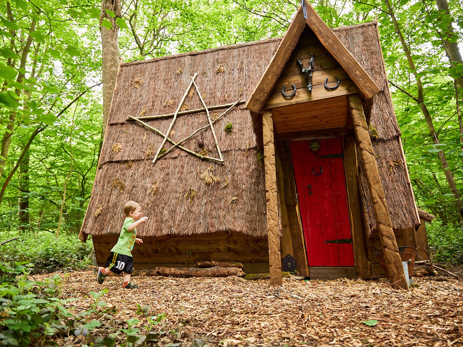 A small child runs toward a whimsical wooden cabin with a red door and a large pentagram made from sticks on the thatched roof, set in a green forest.