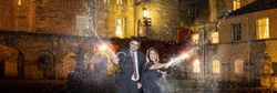 A man and woman celebrating and spraying champagne outside a lit-up stone building at night.