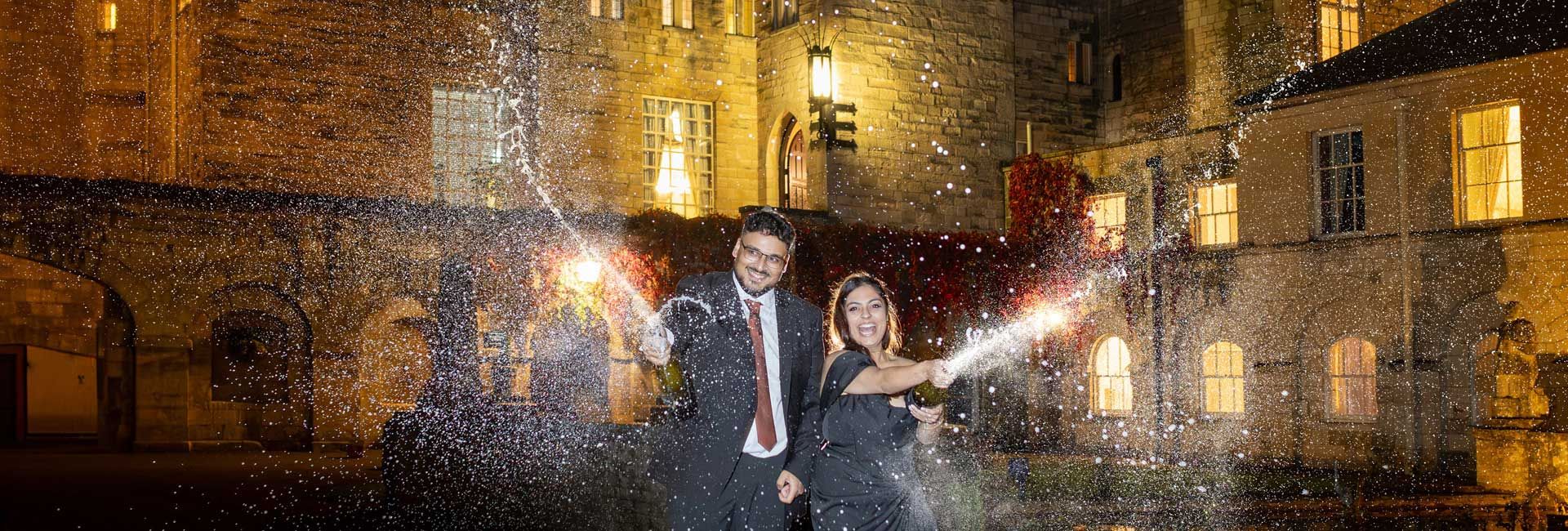 A man and woman celebrating and spraying champagne outside a lit-up stone building at night.
