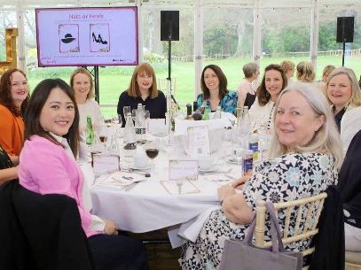 Group of women seated around a round table at a formal lunch event in a glass marquee, with a presentation screen in the background.