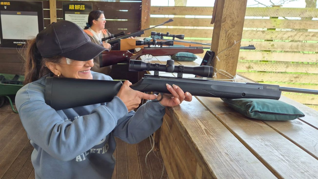 Woman aiming a scoped air rifle at a wooden shooting range with another shooter in the background