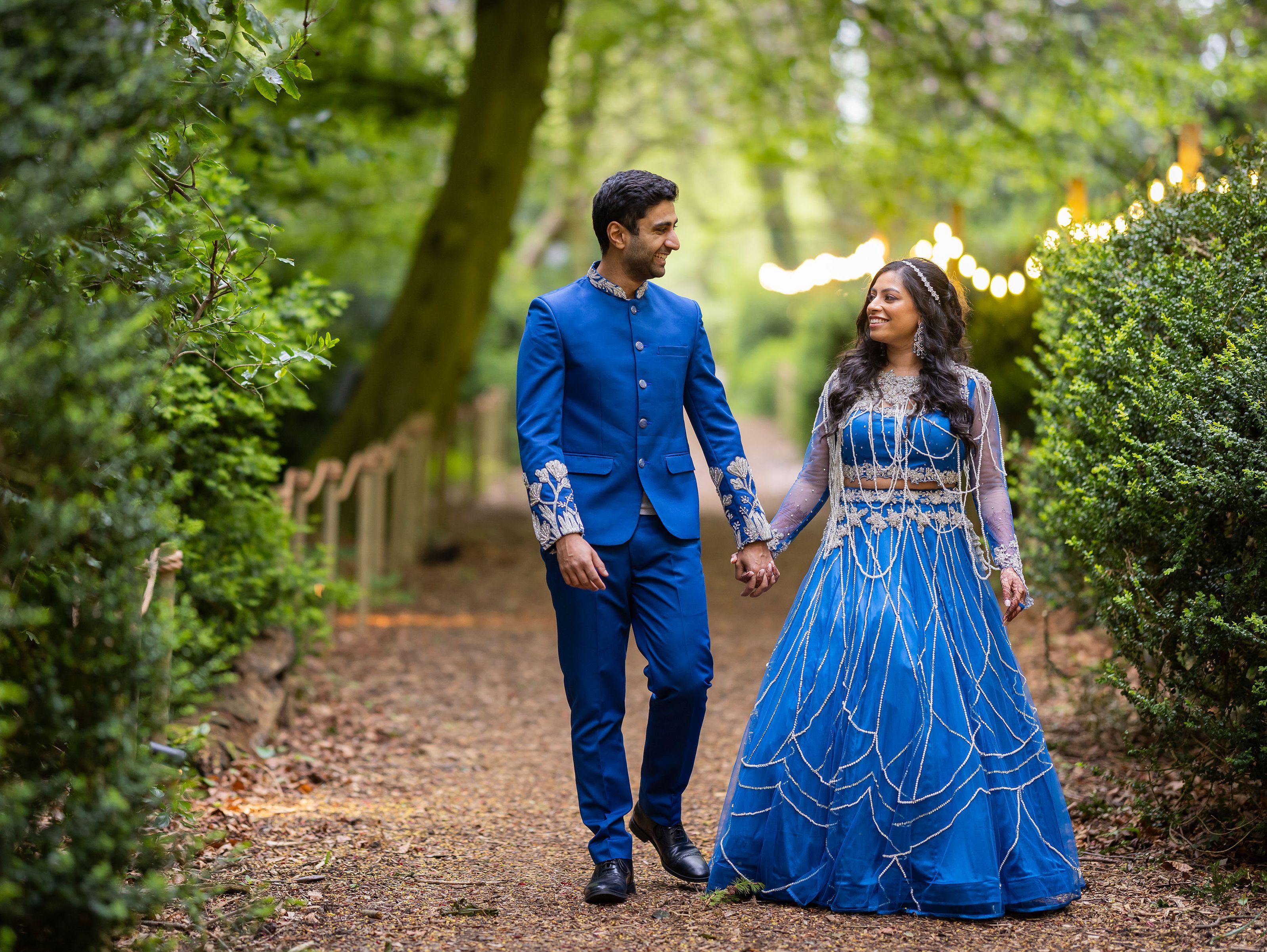 Couple in matching blue traditional outfits holding hands and walking along a woodland path with fairy lights