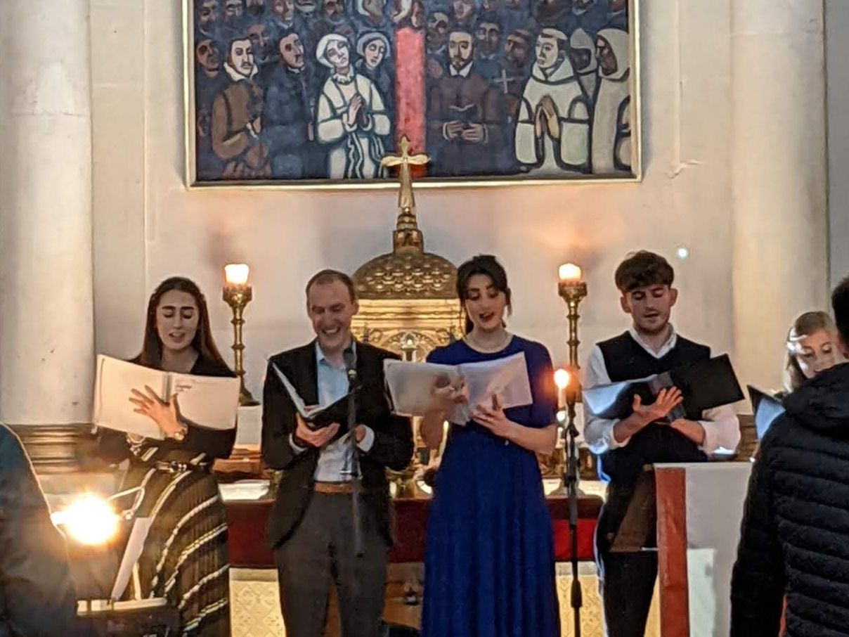 Choir singers performing in a church in front of the altar with candles and hymn books, viewed from the congregation.