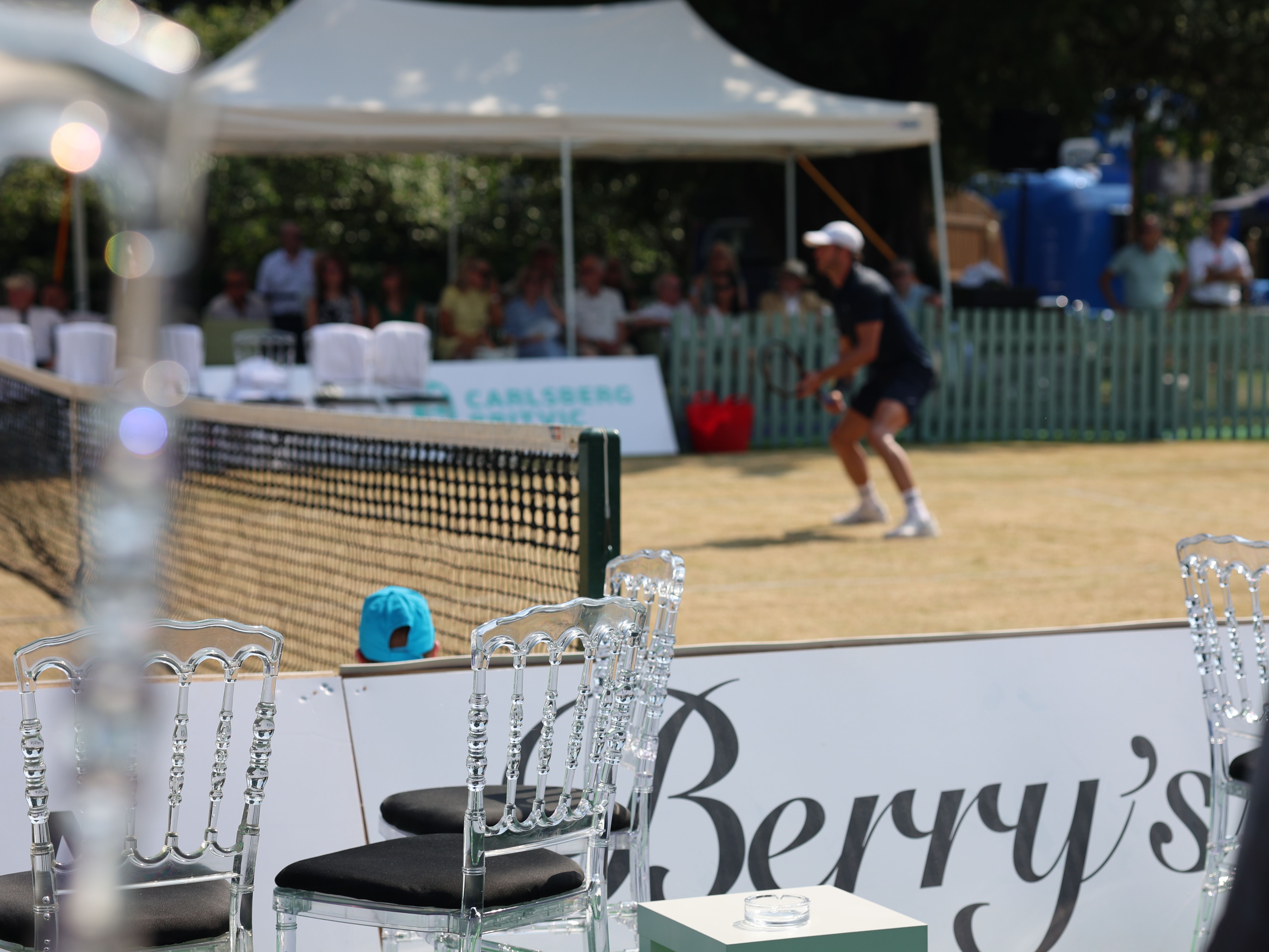 Tennis player preparing to return serve on a grass court with spectators under a white marquee
