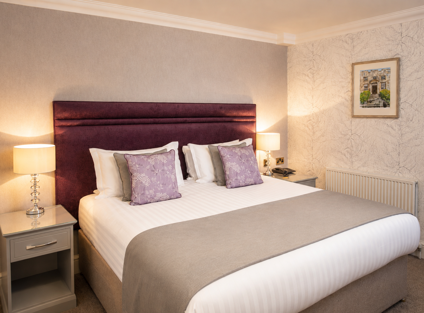 Neatly made double bed with purple headboard, bedside lamps and lavender cushions in a cosy bedroom