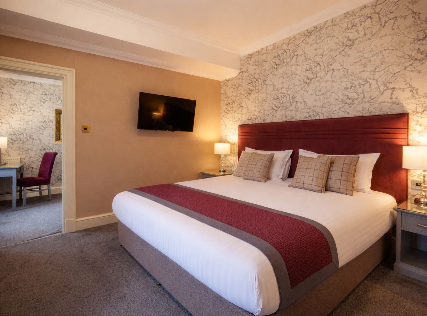 Modern hotel bedroom with king-size bed, red runner, bedside lamps and wall-mounted TV, with an adjoining desk area visible through a doorway.