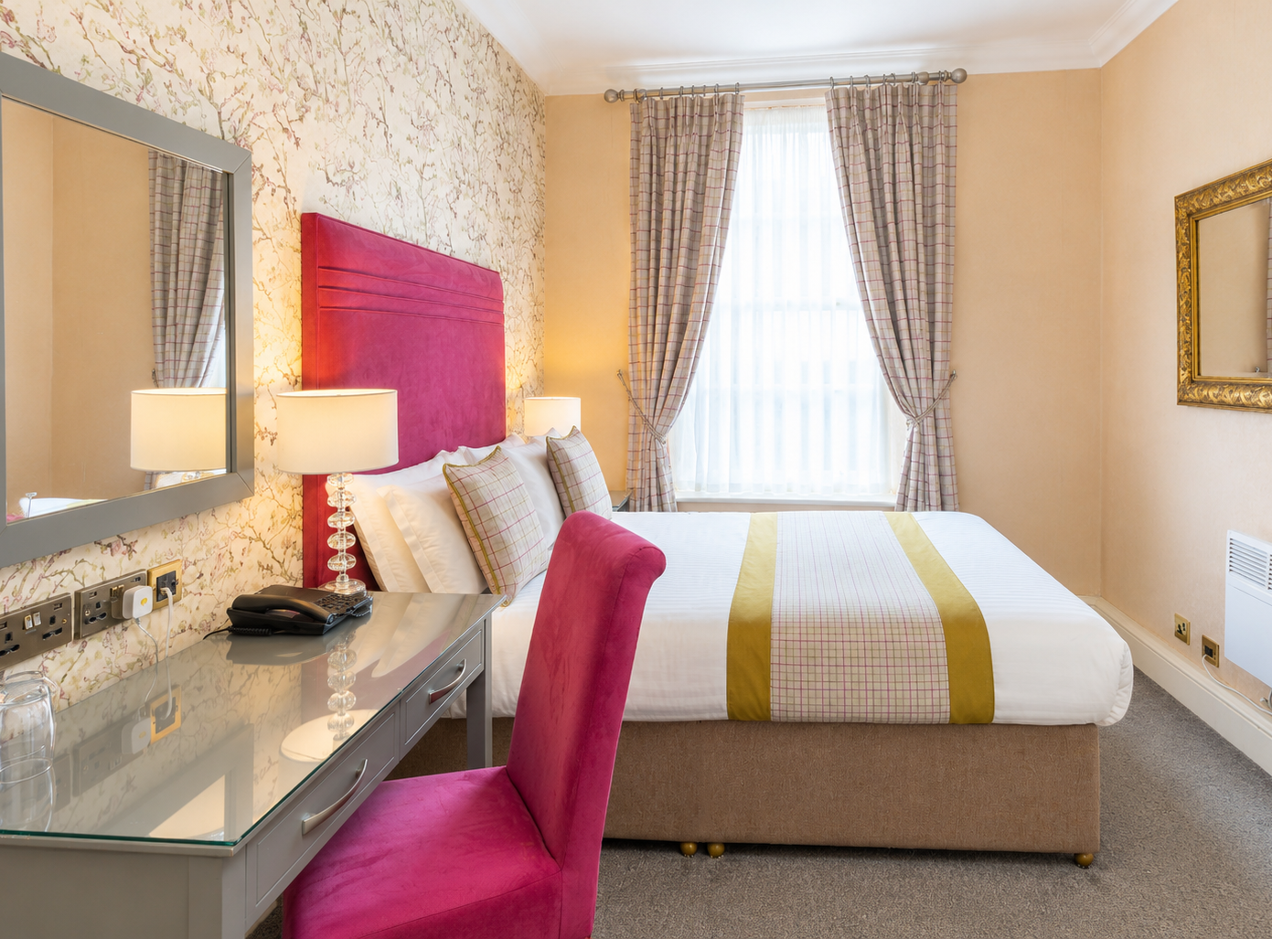Bright hotel bedroom with a pink upholstered headboard, double bed, dressing table and large window with checked curtains