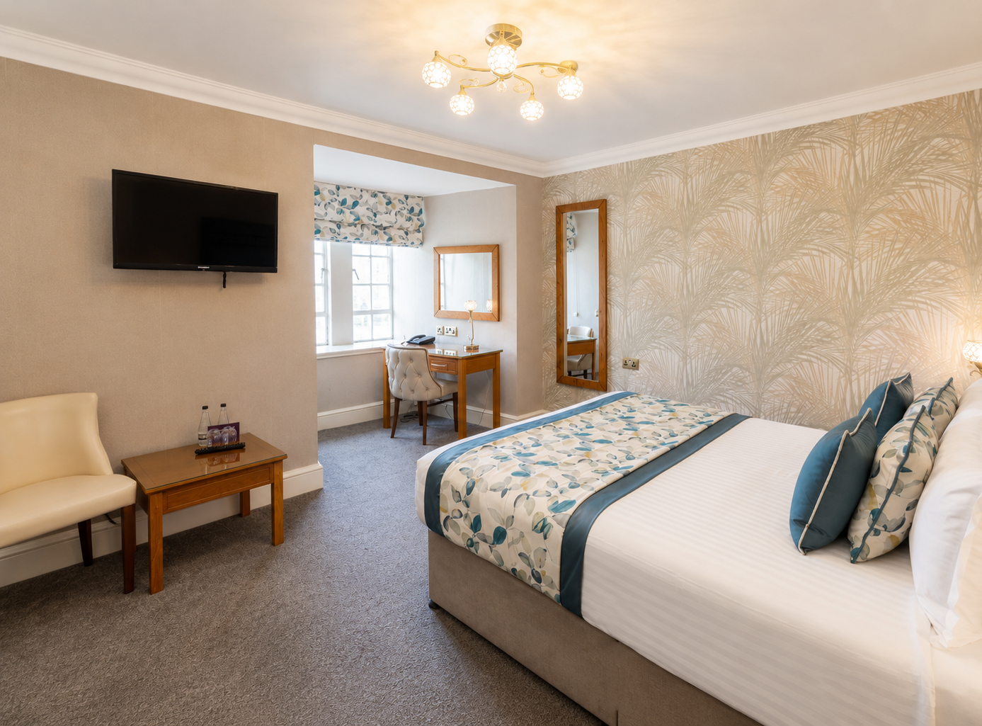 Modern hotel bedroom with double bed, teal cushions, patterned wallpaper, wall-mounted TV and desk by the window