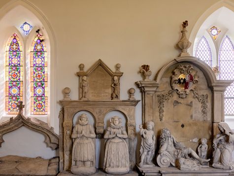 Stone memorial sculptures and plaques inside a church with stained-glass windows