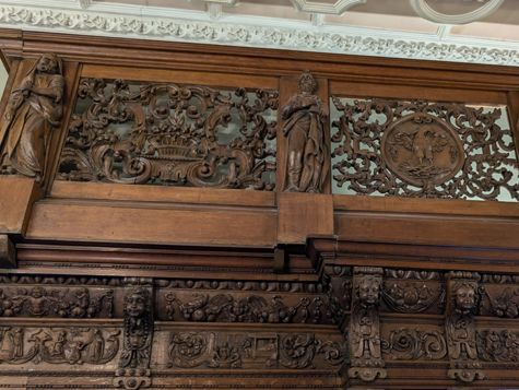 Intricately carved wooden balcony with statues, scrollwork and a decorative crest beneath an ornate ceiling.