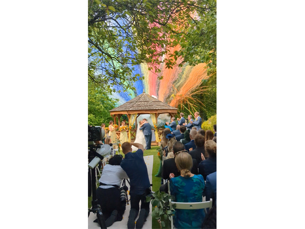 Guests applaud a couple embracing in front of a gazebo at an outdoor wedding ceremony, with colourful fireworks smoke in the sky.