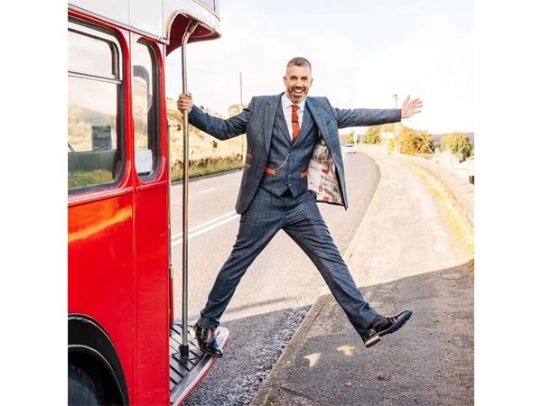 Man in a blue suit stepping off a red bus with arms outstretched on a roadside pavement