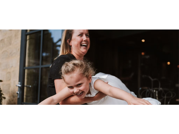 Woman laughing while holding a smiling young girl in a white dress with arms outstretched outside a building
